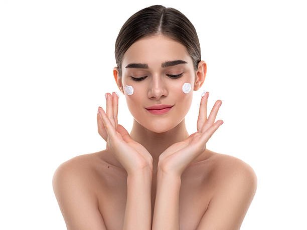 A young woman gracefully applies skincare cream on her cheeks, promoting beauty and healthy skincare. The image conveys a sense of rejuvenation, self-care, and personal wellness routine. Its minimalist background emphasizes the subject's radiant skin and creamy cosmetic application, ideal for skincare, beauty, and wellness-related projects.